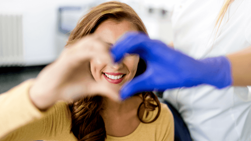 A smiling woman who received emergency dental care in Longmont, CO making a heart shape with her hands and a dentist wearing blue gloves.